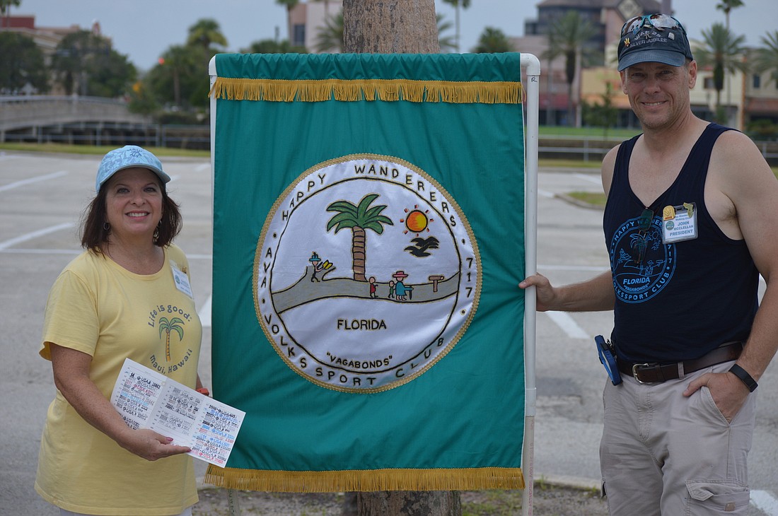 Sheila (left) and John (right) McClellan pose with the Happy Wanderers flag and a card with stamps to note how many walks the pair have been on. Photo by Tim Briggs.