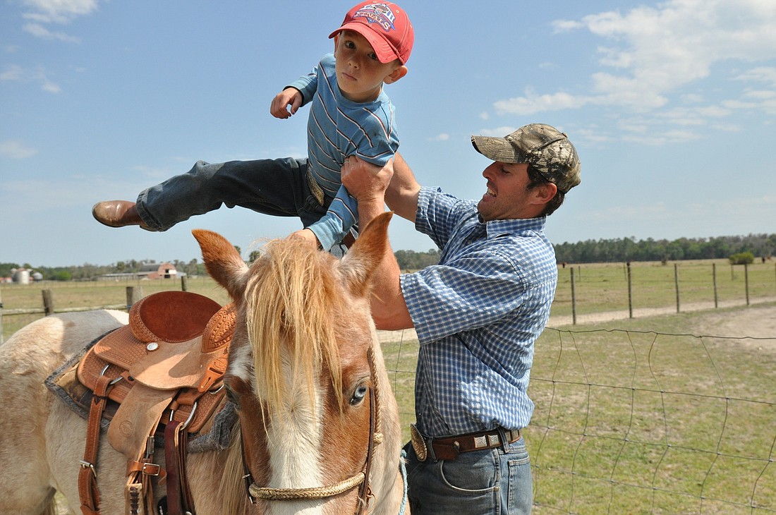 Ramzi Hughes helps his oldest son Ramzi on a horse before rounding up cattle to practice steer roping for Cracker Day.