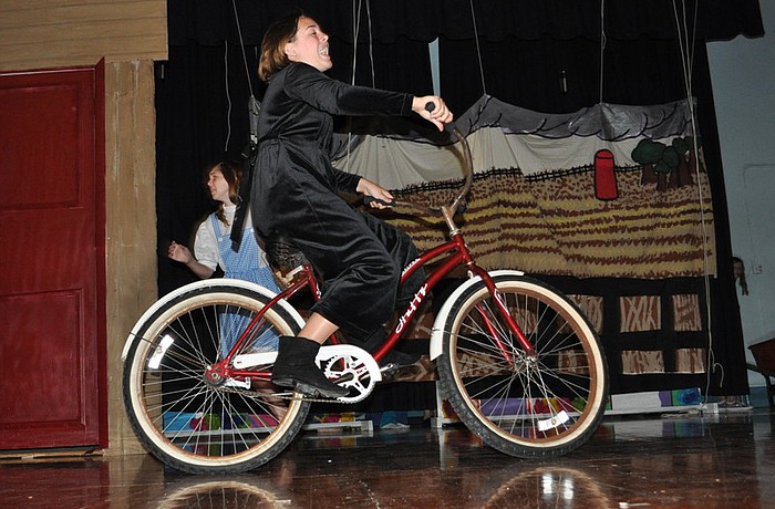 Caught up in the tornado, Brianna McVaugh, acting as Miss Almira Gulch, rides her bicycle across the stage.