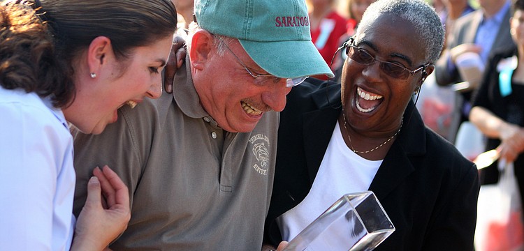 County Commissioners Christine Robinson and Carolyn Mason smile and laugh as they surrounded a teary Norman Schimmel after he was surprised during the 2011 National Tourism Week Awards ceremony and given the â€œVoice of Tourismâ€ Award.