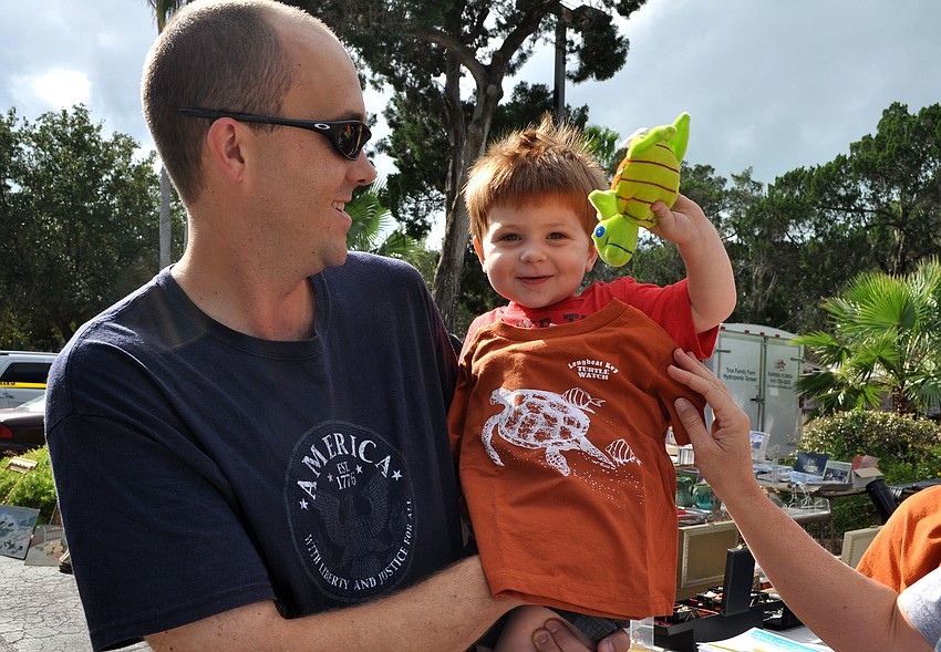 Cal Leggett holds his son, Alex, 2, as he gets sized for a Longboat Key Turtle Watch shirt and holds up a stuffed toy turtle Sunday, Oct. 28, during the Fall Festival at the Centre Shops.