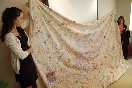 Downtown Vision Inc. Communications Coordinator Sarah Henderson and DVI Marketing and Events Assistant Liz Grebe show DVI's board of directors the banner that was signed March 7 by hundreds of people at the 100th First Wednesday Art Walk.