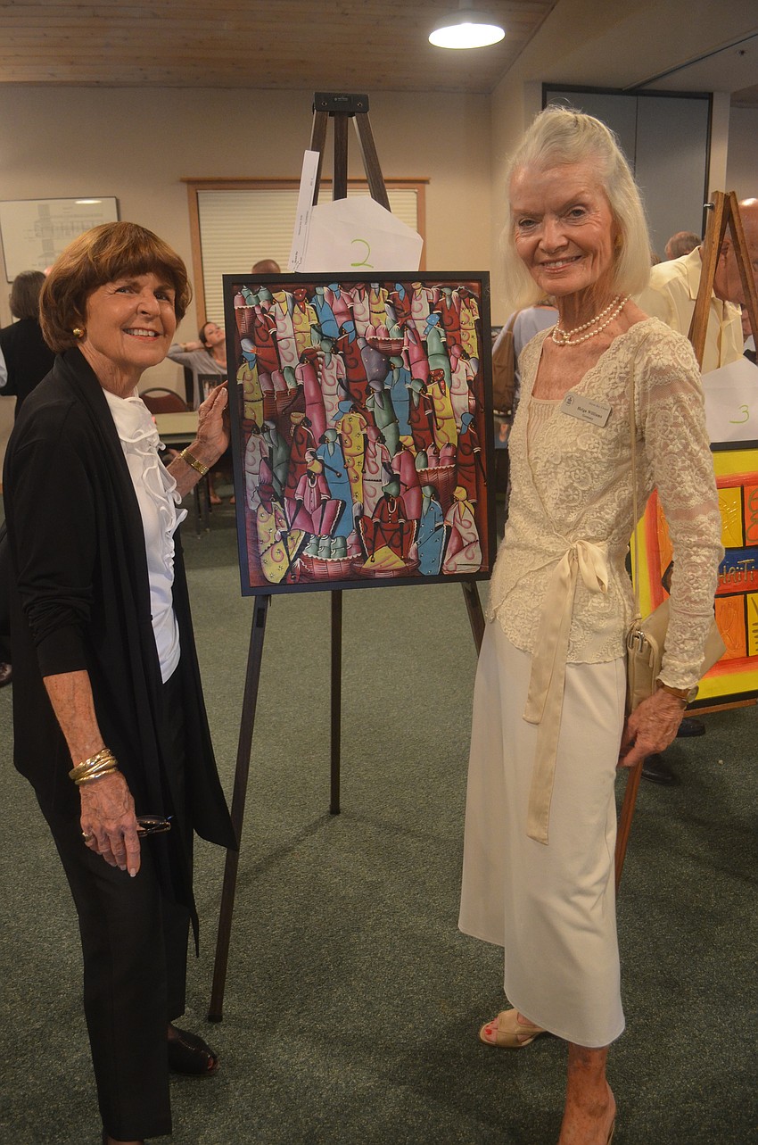 Betsy McLean and Helga Williams display a Haitian painting that is part of the silent auction.