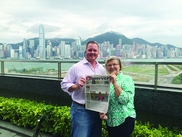 Michael Gibson surprised his mom, Kristina Gibson, and dad, not pictured, as they disembarked in Hong Kong on one of their stops on a Queen Mary 2 world cruise. They read their Observer while overlooking the Central District of Hong Kong from Kowloon.