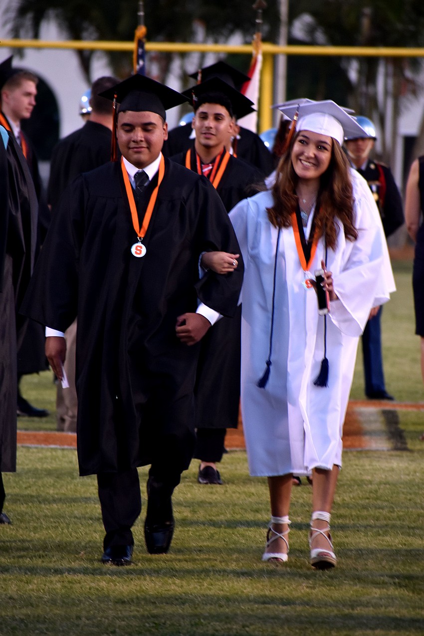 Graduates smile and wave to friends and teachers as they walk to their seats.