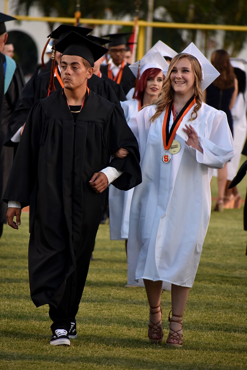 Graduates smile and wave to friends and teachers as they walk to their seats.