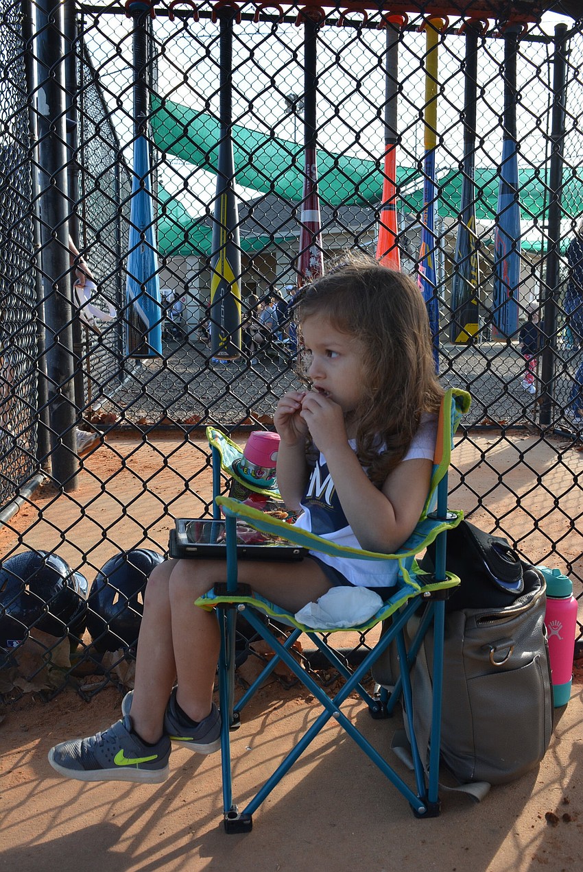 Three-year-old Hadley Drohan has a prime dugout spot for snacking and watching her brother, Jefferson, play in his first game of the season.
