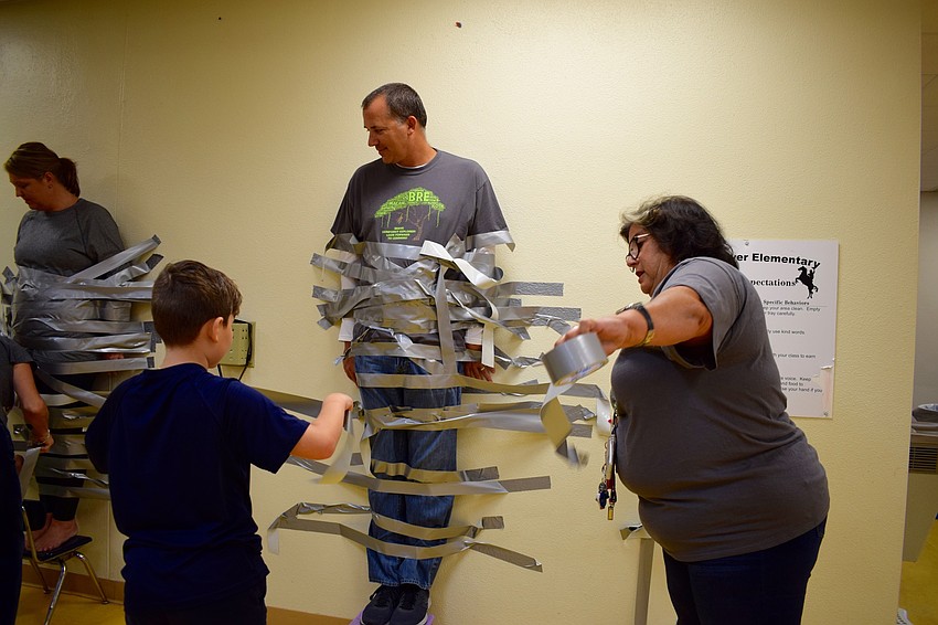 Braden River Elementary School fifth-grader Jonathan Lopez, 10, left, goes to tape Principal Joshua Bennett, center, to the wall while ESOL paraprofessional Barbara Grimes, right, rips off more tape to hand to students.