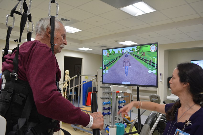 Physical therapist Dr. Julia Figueroa (right) points out the objective of a Hocoma Lokomat-based game to stroke patient Ronald Serres. In this game, Serres must walk a certain speed to collect coins before a CPU opponent.