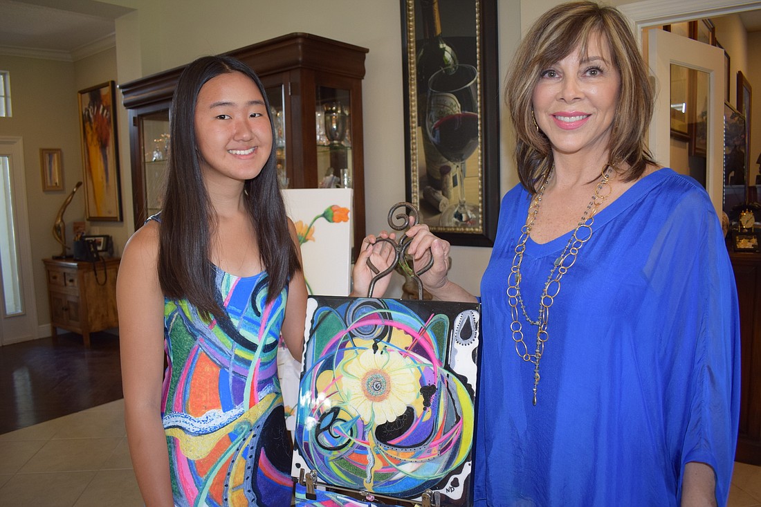Tiffany Yu wears a dress by Debbie Dannheisser Threads while Debbie Dannheisser shows a painting by her late daughter Nichole. She incorporates Nichole&#39;s artistry into her fashions.