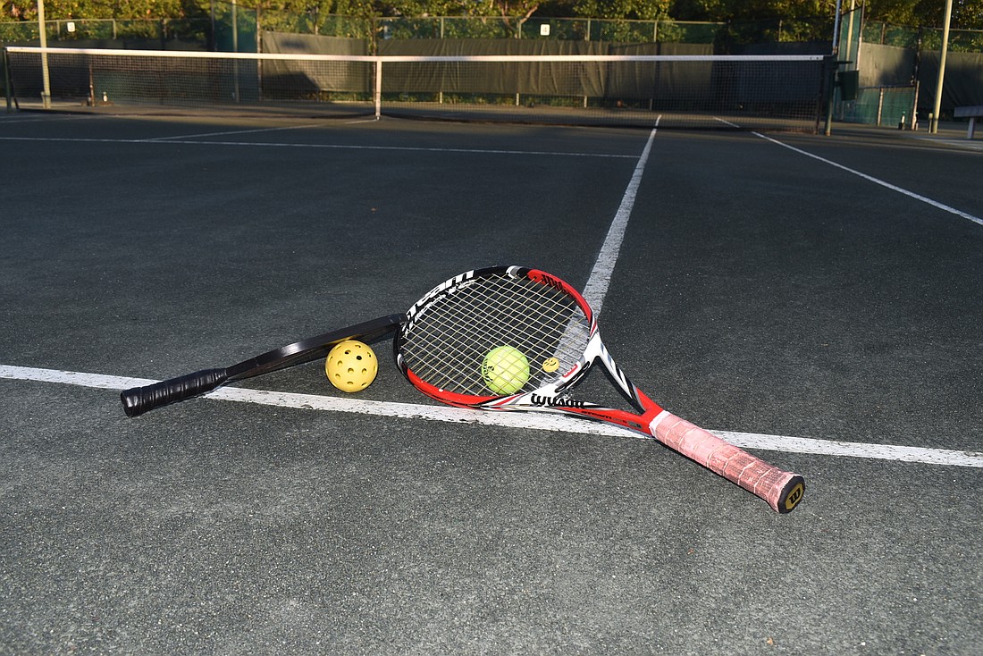 A tennis racket and ball and a pickleball racket and ball lie on a court at Cedars Tennis Resort & Club. Although the two sports are related, they have some differences in health benefits and drawbacks.