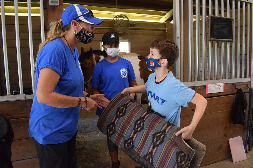 Samantha Toomey, equine manager, and volunteer Ellie Blitz, help 10-year-old camper Joseph Woolbert put a saddle pad on Buddy Cassidy.