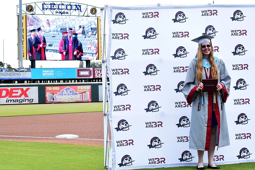 Graduates continue to cross the baseball diamond while Madigan Wilford gets her photo taken with her diploma.