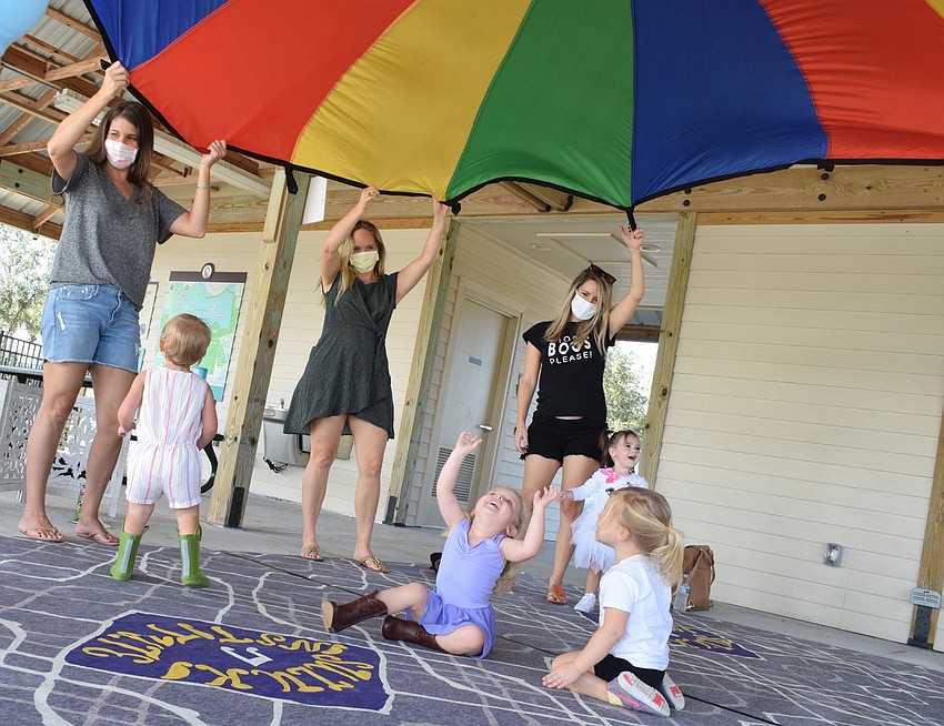 The moms throw up the parachute as the children wait for it to fall on top of them.