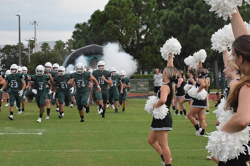 Football players rush the field to start the game as cheerleaders cheer for them.