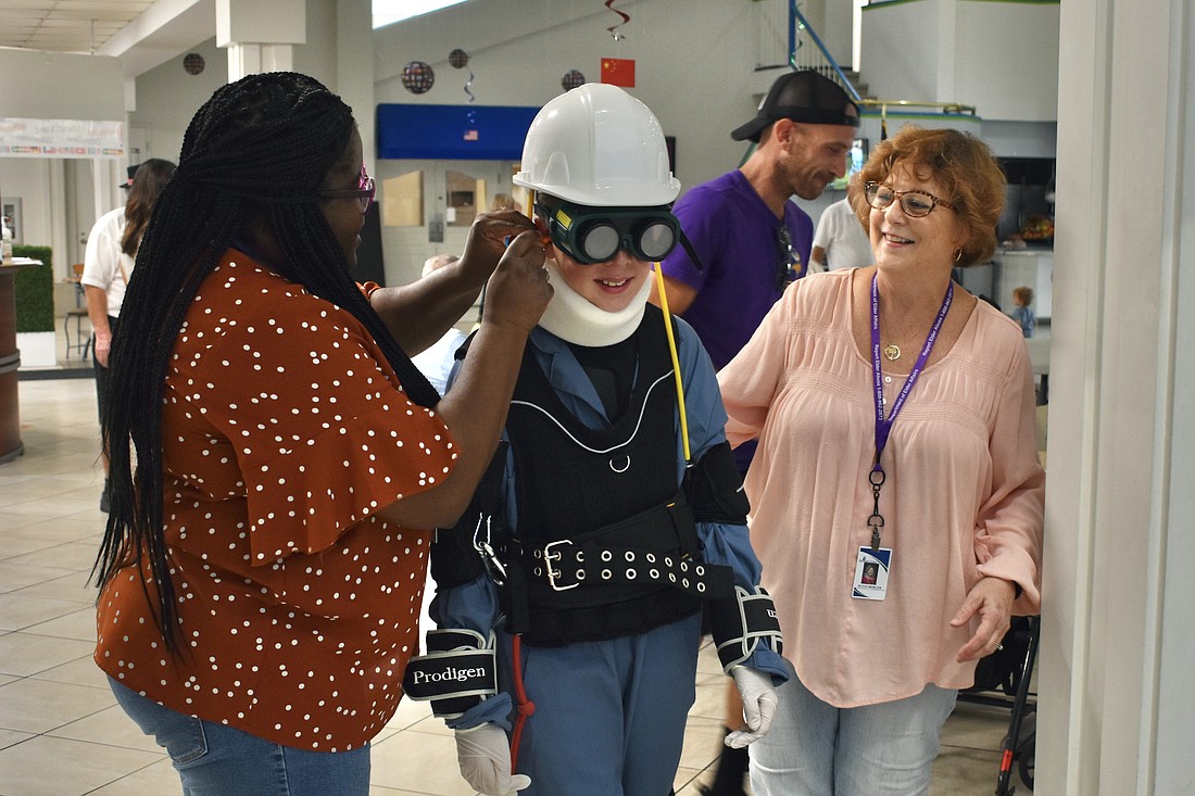 Florida Department of Health employee Quiana Tolbert helps 11-year-old Beckett Hill into the Sarasota Aging Sensitivity Suit, alongside Aging System Policy Coordinator Susan Berger.