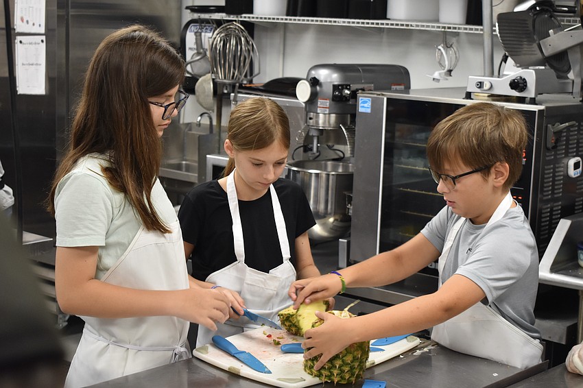 Eleven-year-old Emma Granthon, and 12-year-old Abby Williams and Asher Woods work together to slice a pineapple.