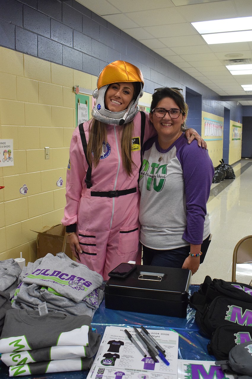 Parents Hannah O'Reilly and Amanda Hasenzahl are ready to sell school merchandise to boost school spirit. O'Reilly dresses as an astronaut to match the space theme of the book fair.