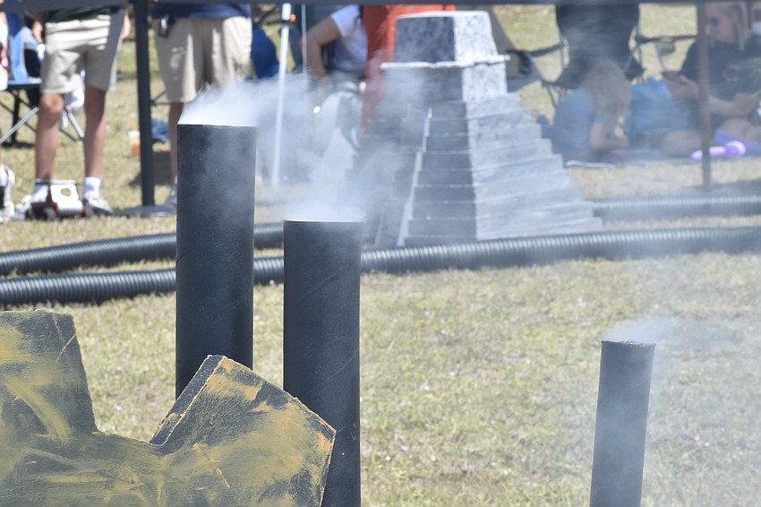 Smoke rises from smokestacks in the industrial revolution, in front of an Aztec pyramid on the other side of the track.