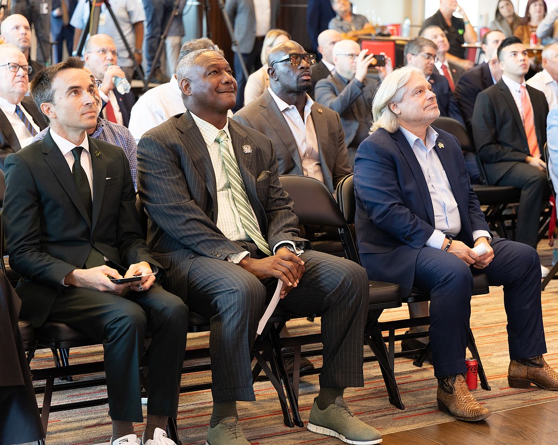 Rob Higgins, CEO of USF Athletics, Derrick Brooks, former Tampa Bay Buccaneer and NFL and college football Hall of Famer, and Santiago Corrada, CEO, Visit Tampa Bay watch a presentation on the 2029 CFP championship game coming to Tampa. Titus O’Neil, a retired professional wrestler and college football player at the University of Florida looks on from the second row.