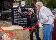 With scissors in one hand and a whip in the other, Commissioner Carol Felts cuts the ribbon on a historical marker for The Florida Cracker Trail Feb. 13 at Manatee Village Historical Park.