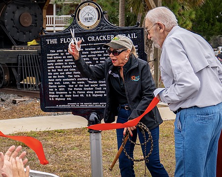 With scissors in one hand and a whip in the other, Commissioner Carol Felts cuts the ribbon on a historical marker for The Florida Cracker Trail Feb. 13 at Manatee Village Historical Park.