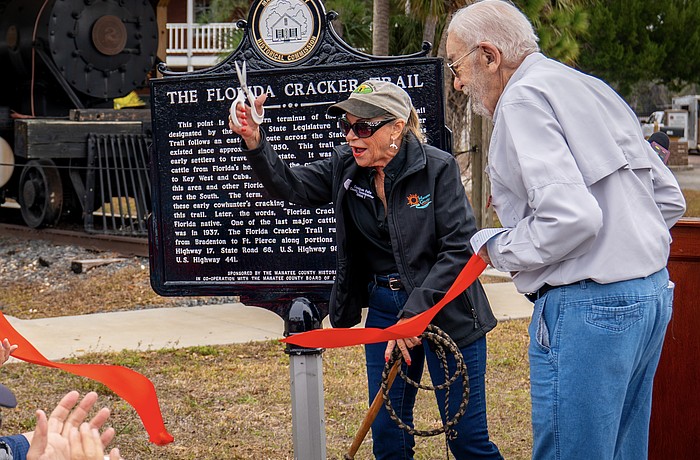 With scissors in one hand and a whip in the other, Commissioner Carol Felts cuts the ribbon on a historical marker for The Florida Cracker Trail Feb. 13 at Manatee Village Historical Park.