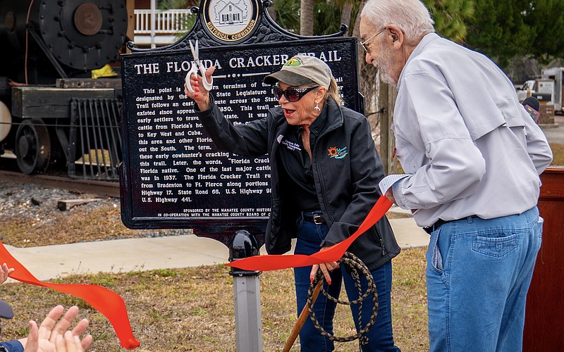 With scissors in one hand and a whip in the other, Commissioner Carol Felts cuts the ribbon on a historical marker for The Florida Cracker Trail Feb. 13 at Manatee Village Historical Park.
