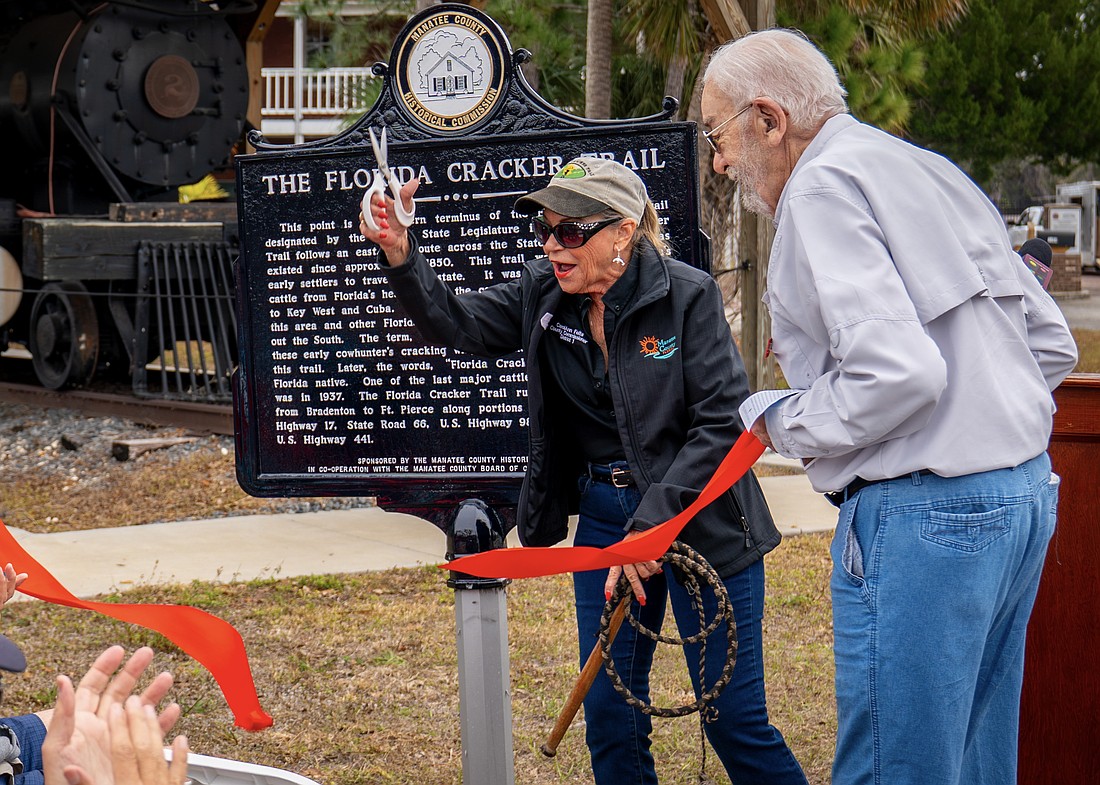With scissors in one hand and a whip in the other, Commissioner Carol Felts cuts the ribbon on a historical marker for The Florida Cracker Trail Feb. 13 at Manatee Village Historical Park.